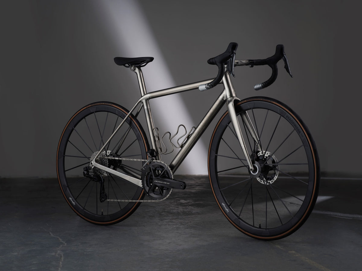 Titanium road bike with tan-wall tyres and titanium bottle cages under a diagonal light beam