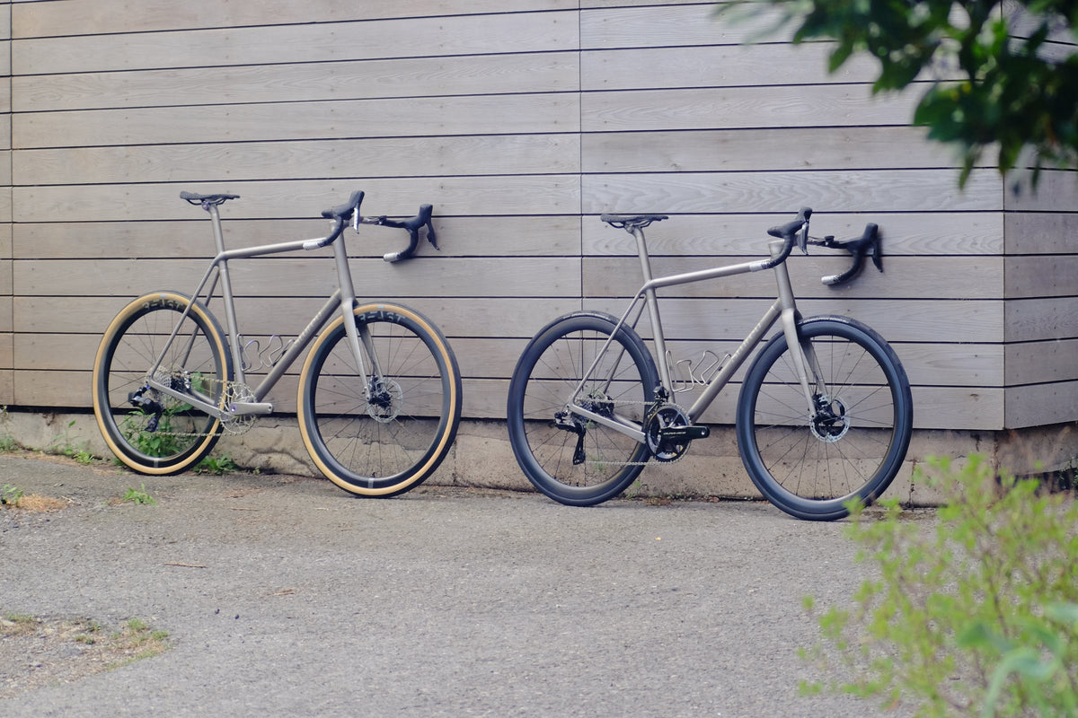 Two titanium road bikes, one with tan-wall tyres, leaning against a grey timber-clad wall