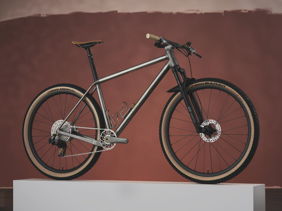 Titanium hardtail mountain bike with tan-wall tyres and suspension fork on a white plinth, red backdrop
