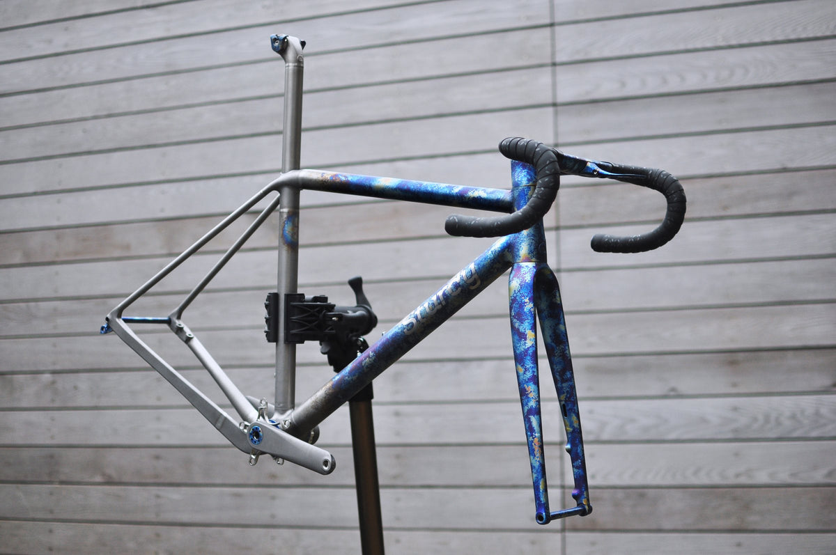 Titanium frame and fork with mottled blue heat-tint finish and integrated bar on a workstand by timber cladding