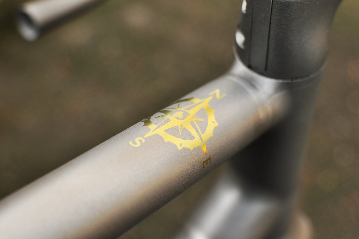 Gold compass rose motif on a shot-peened titanium top tube beside the head tube