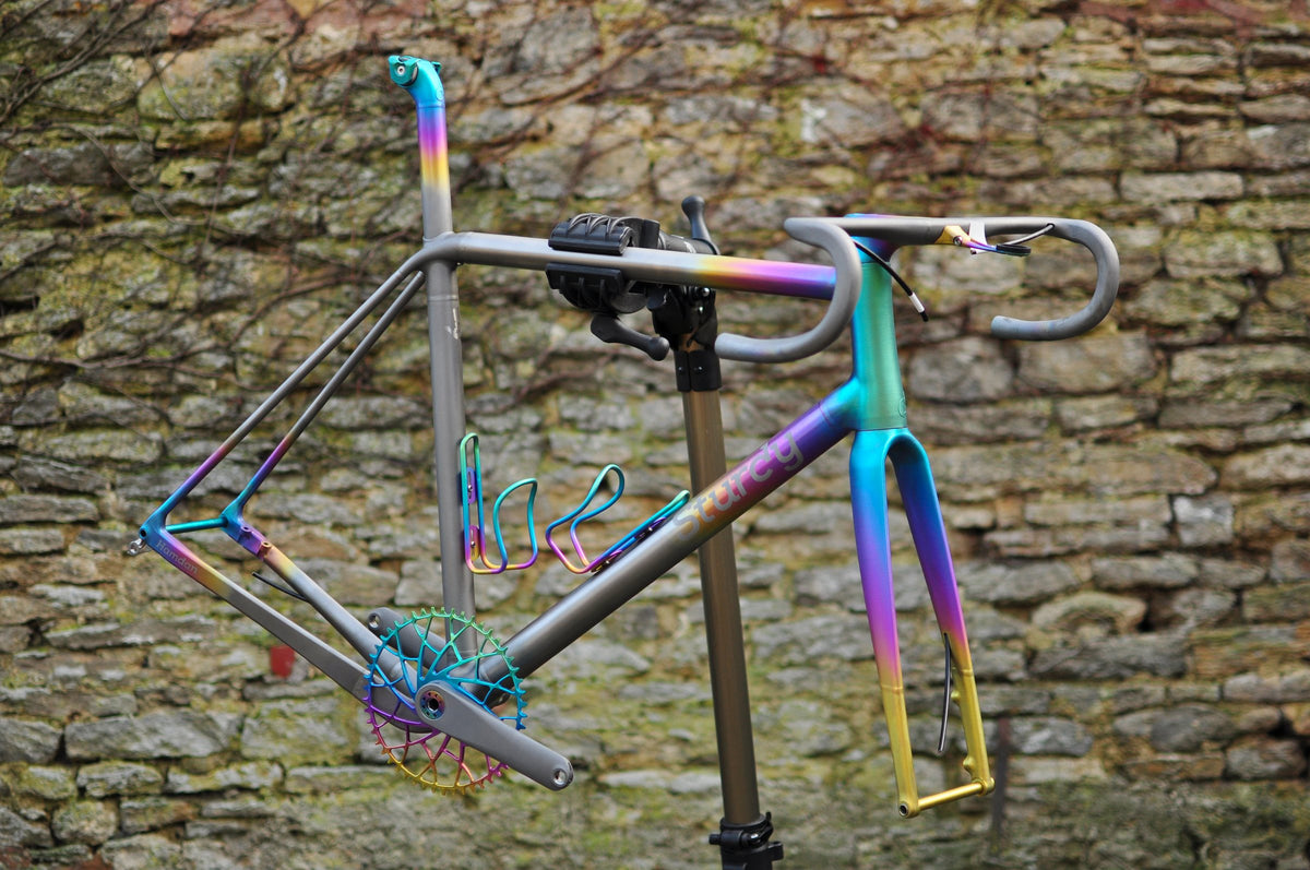 Titanium road frame with rainbow anodised fade and matching bottle cages on a workstand by a stone wall