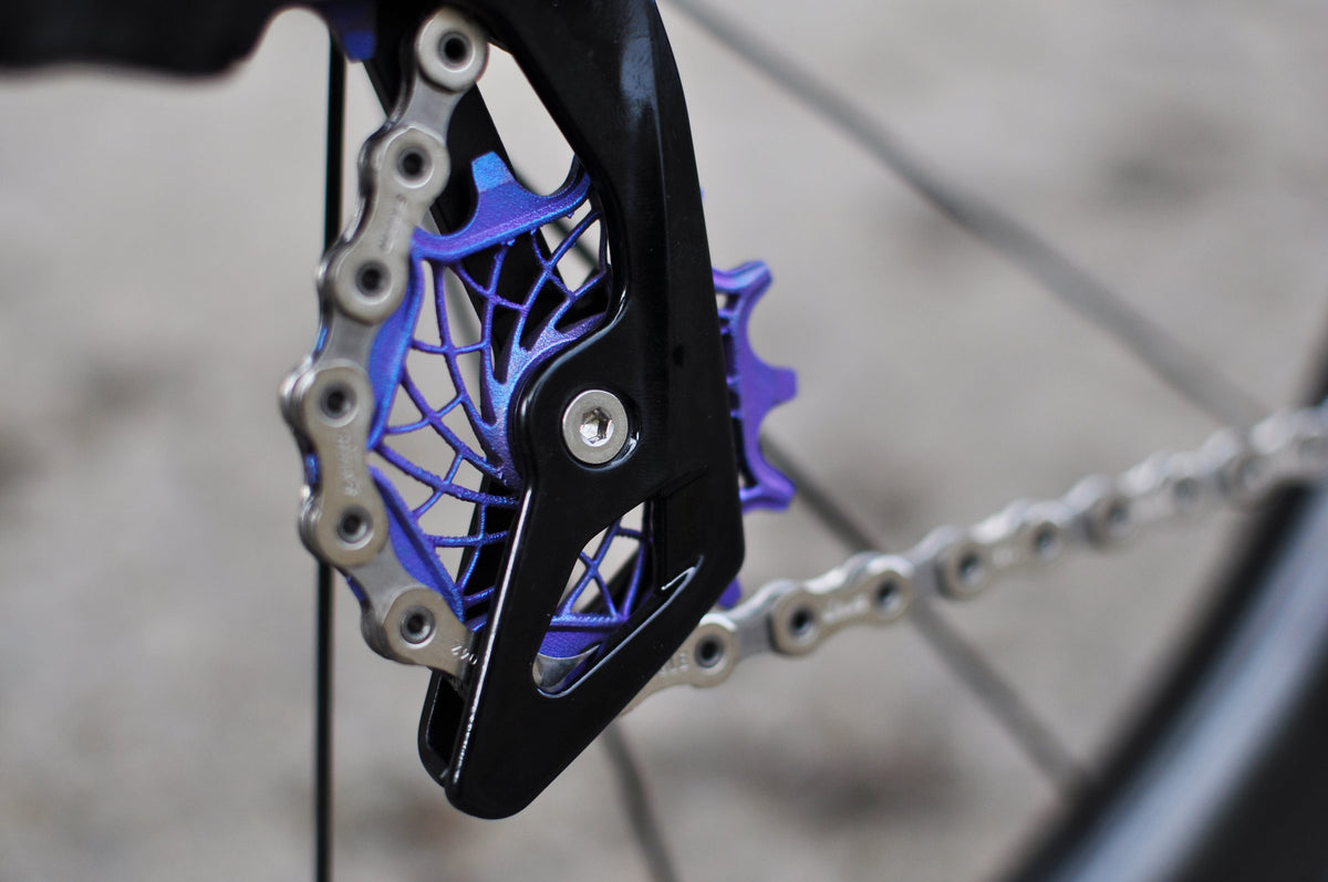 Rear derailleur cage with purple anodised web-pattern jockey wheels and chain, close-up