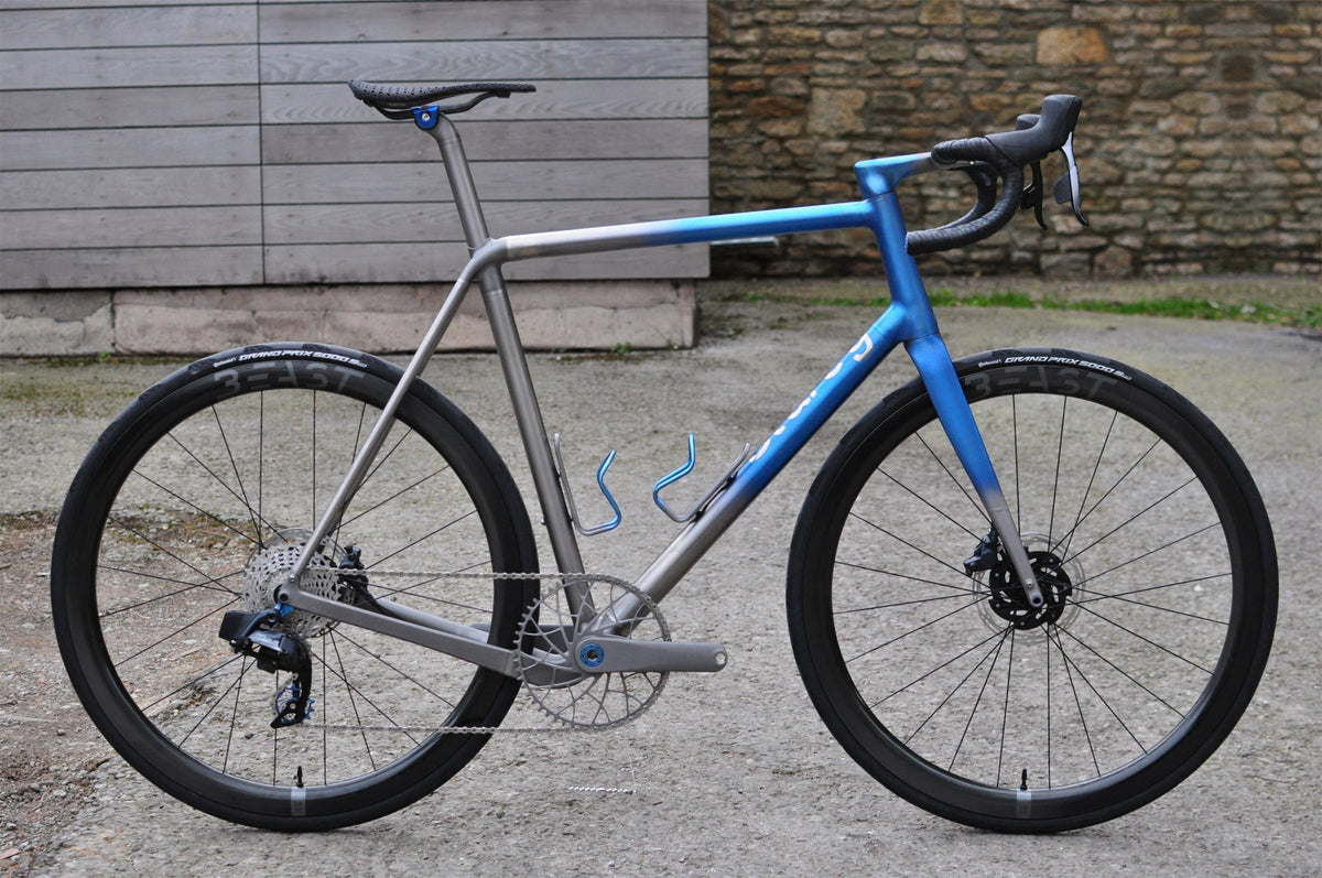 Titanium road bike with blue fade on the front triangle and fork, blue bottle cages, beside a stone wall