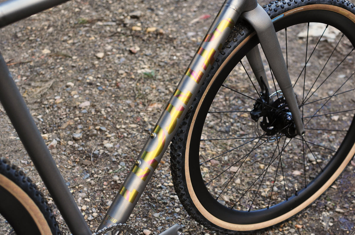 Titanium down tube with yellow and pink anodised lettering beside a tan-wall gravel tyre