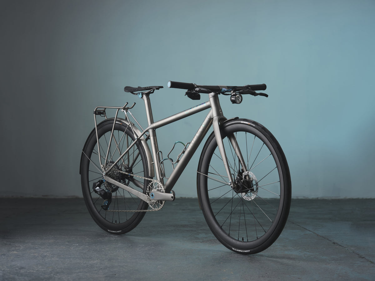 Titanium flat-bar bike with rear rack, mudguards, front light and dark wheels in a blue-lit studio