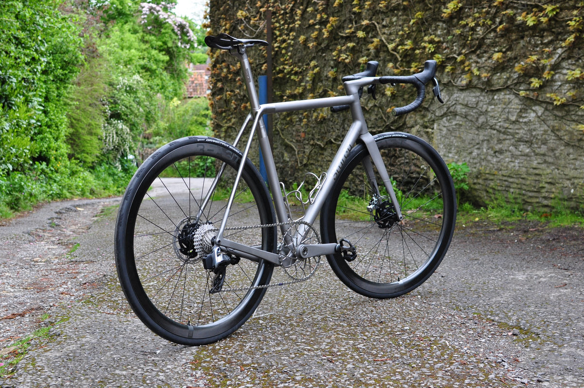 Raw titanium road bike on a gravel lane between a green hedge and an ivy-clad stone wall