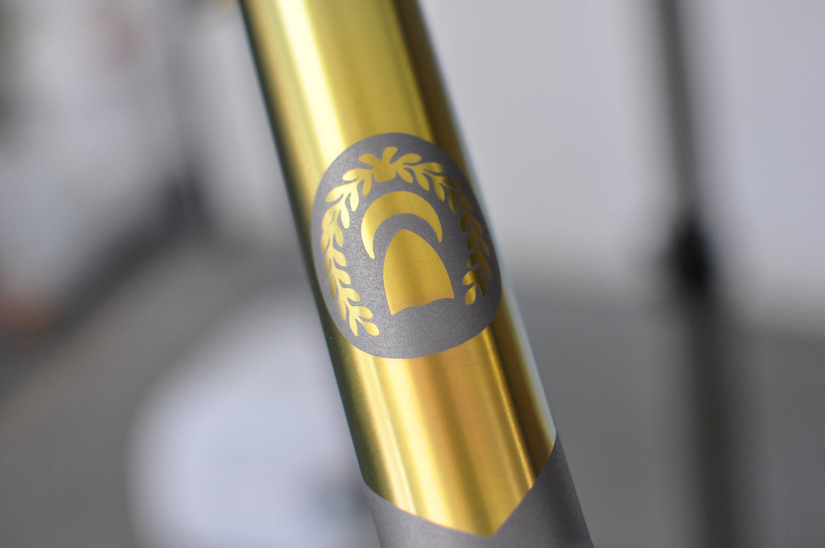 Gold anodised tube with a laurel and arch emblem masked out in shot-peened titanium