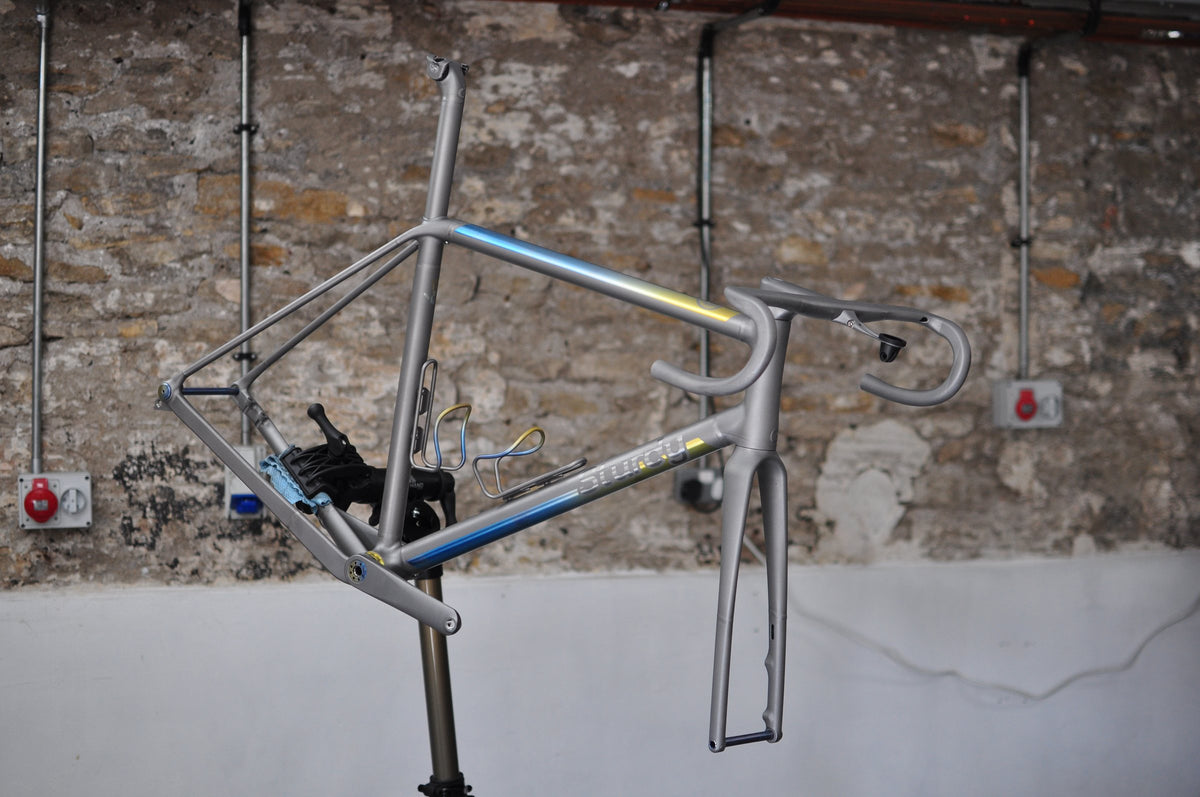 Titanium frame with a blue-to-gold anodised fade on the tubes, on a workstand in a stone-walled workshop