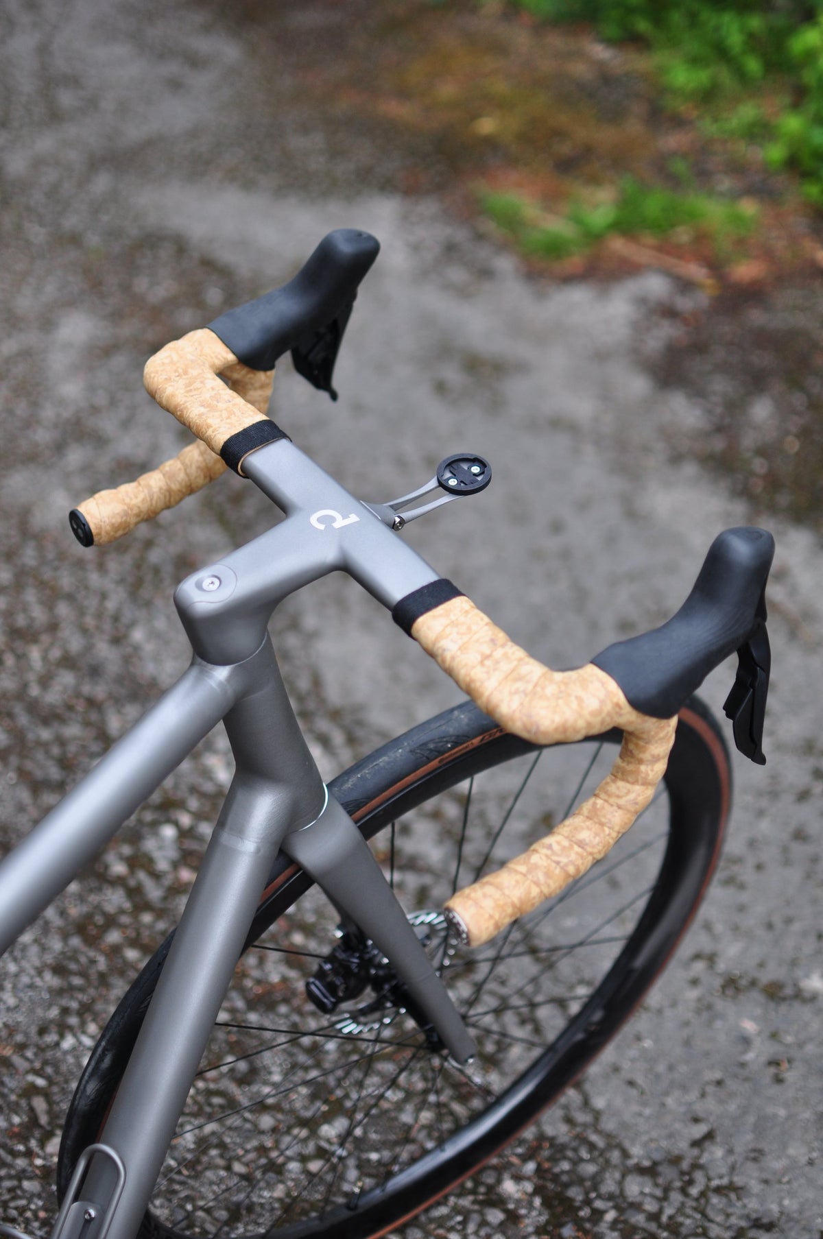 Front end of a titanium road bike with cork-effect bar tape and integrated handlebar on a wet lane