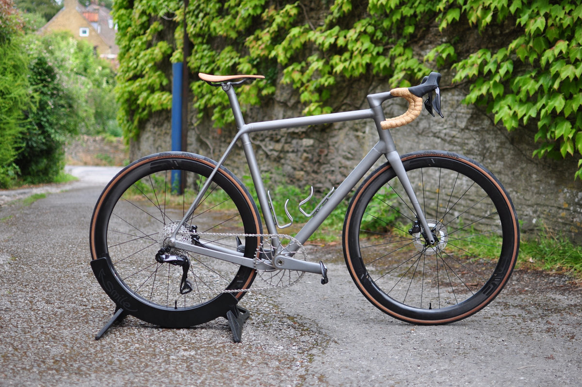Titanium road bike with tan saddle, cork-effect bar tape and tan-wall tyres on a stand by an ivy wall