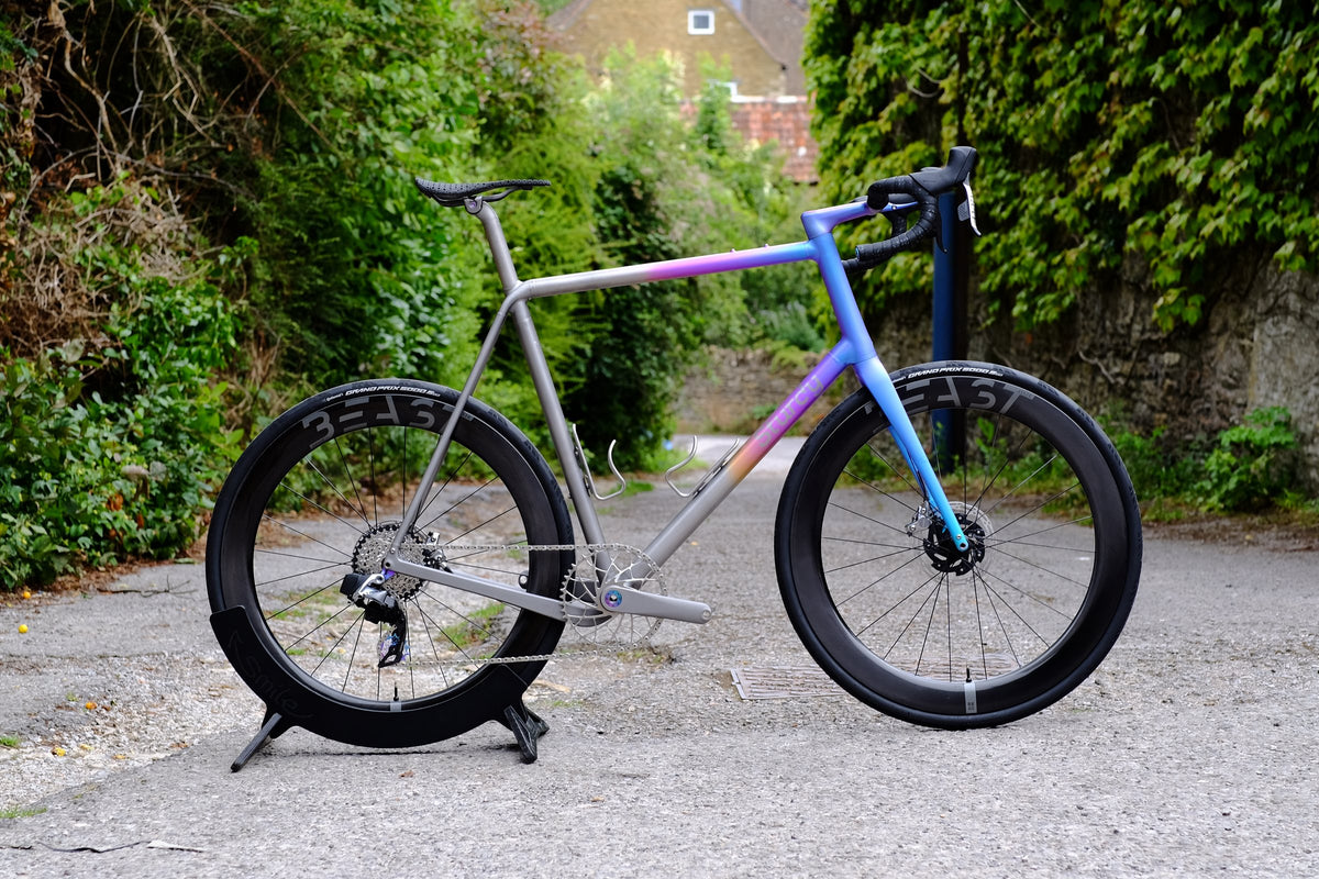Titanium road bike with purple-to-blue fade front end and deep carbon wheels in a leafy lane