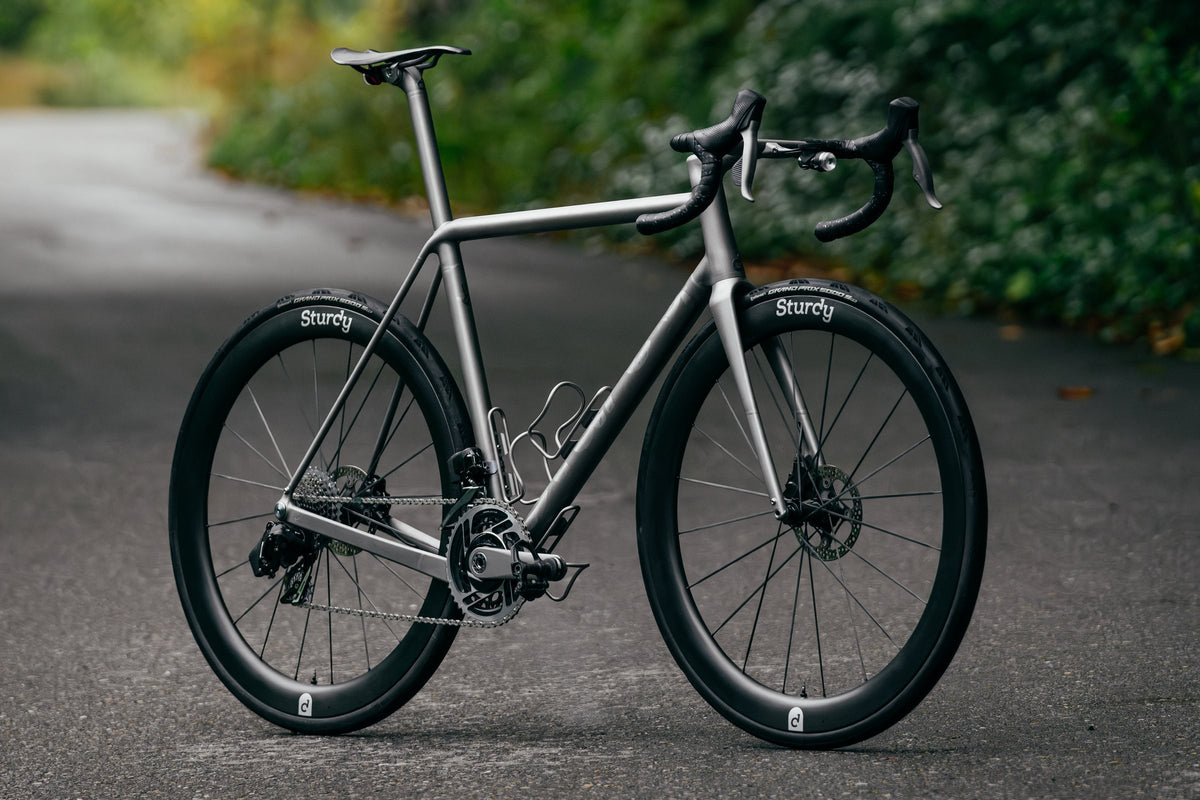 Titanium road bike with Sturdy-branded deep-section wheels on a tree-lined tarmac lane