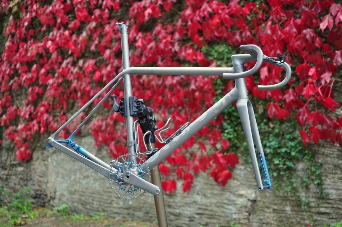 Titanium road frameset with blue anodised details and integrated bar in a stand before red ivy