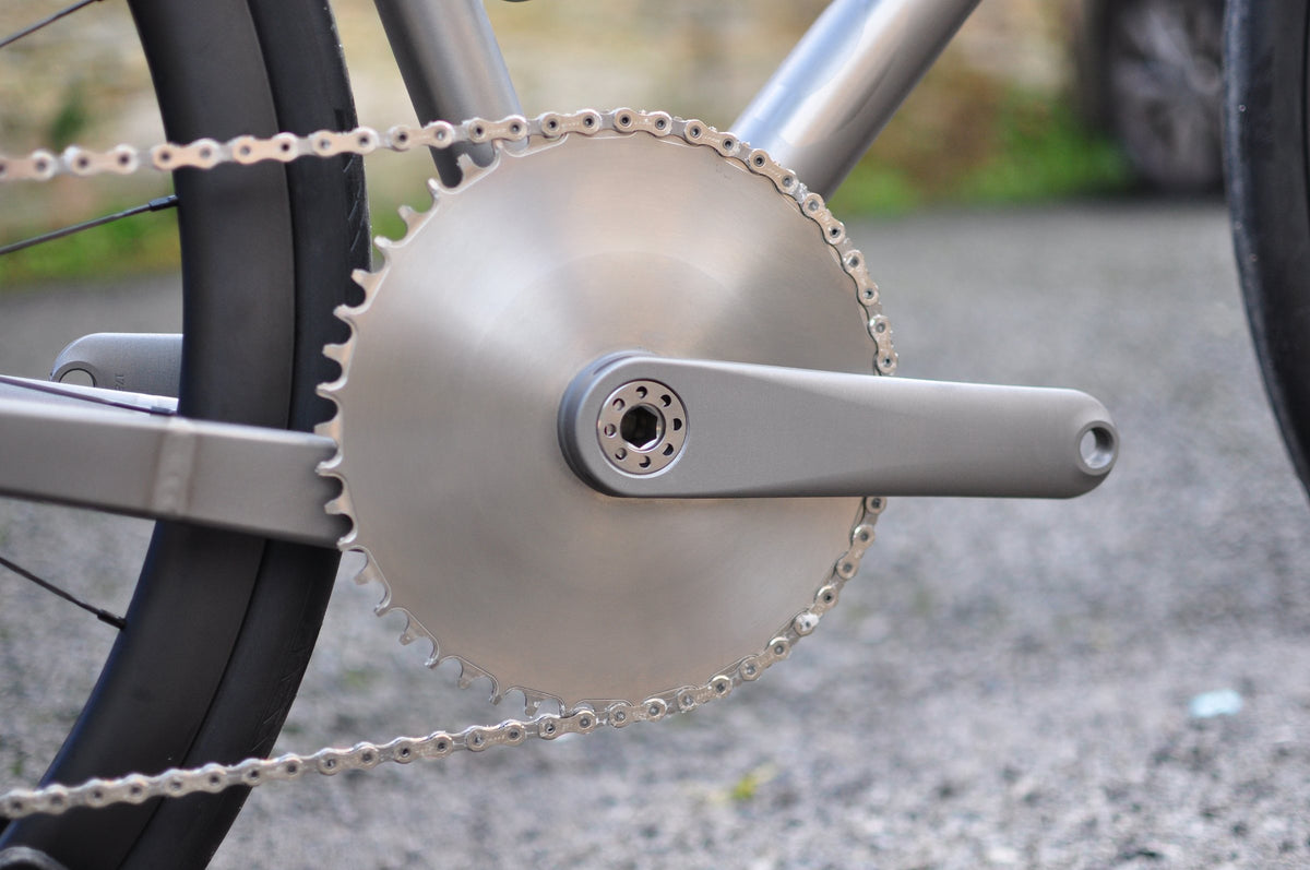 Brushed titanium disc chainring and titanium crank arm with chain, gravel courtyard behind