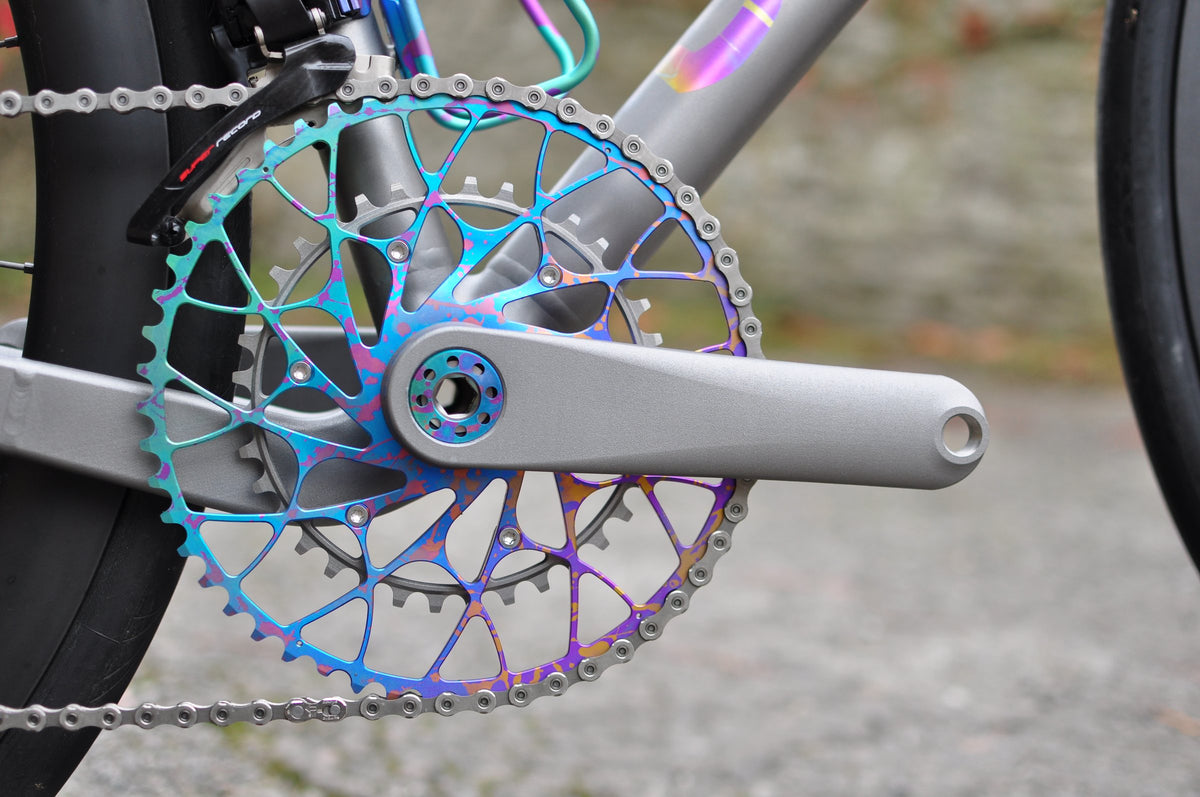 Rainbow anodised titanium skeletal chainring with grey crank arm and anodised bottle cage behind