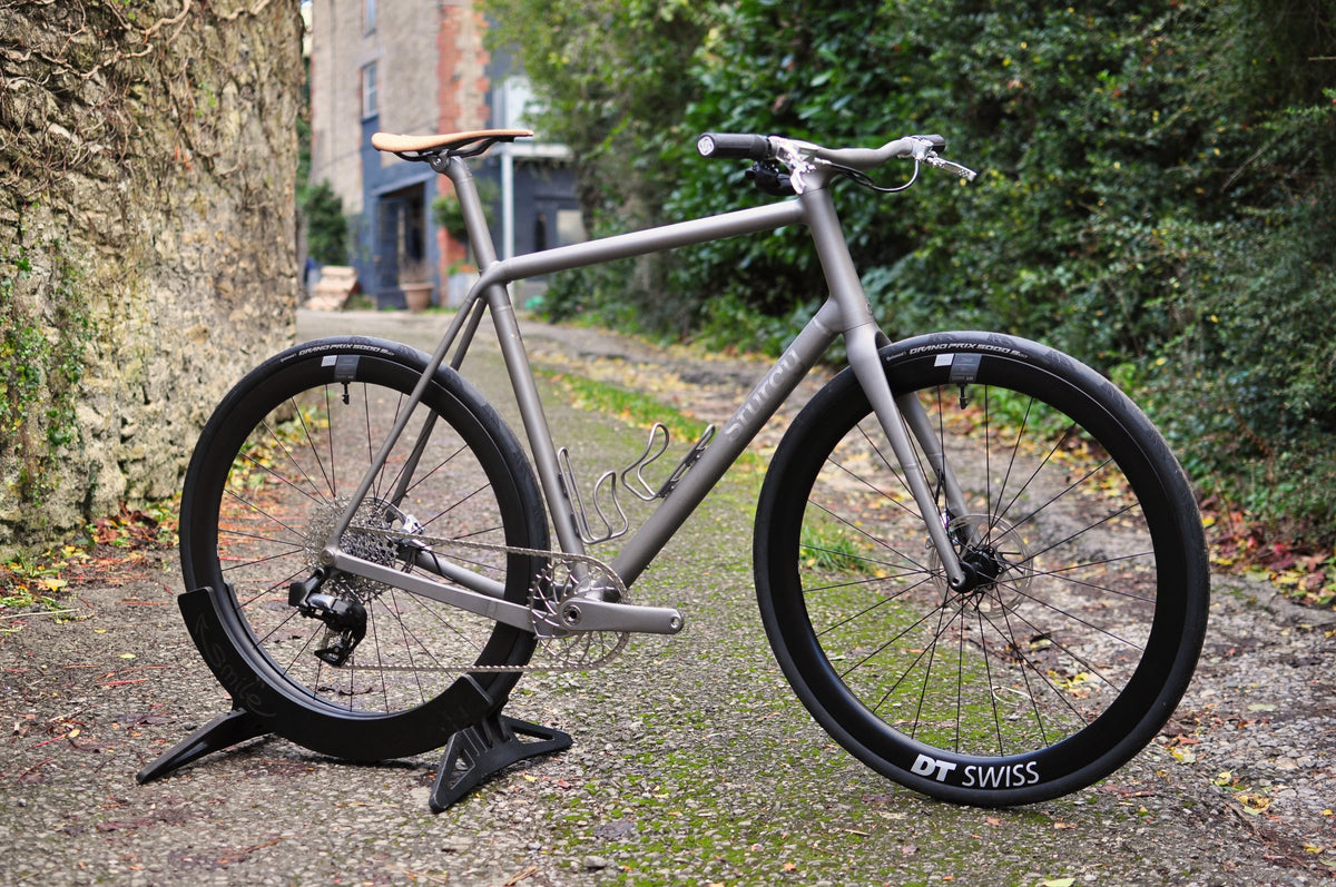 Titanium flat-bar bike with tan saddle and deep-section wheels on a stand in a mossy lane by a stone wall