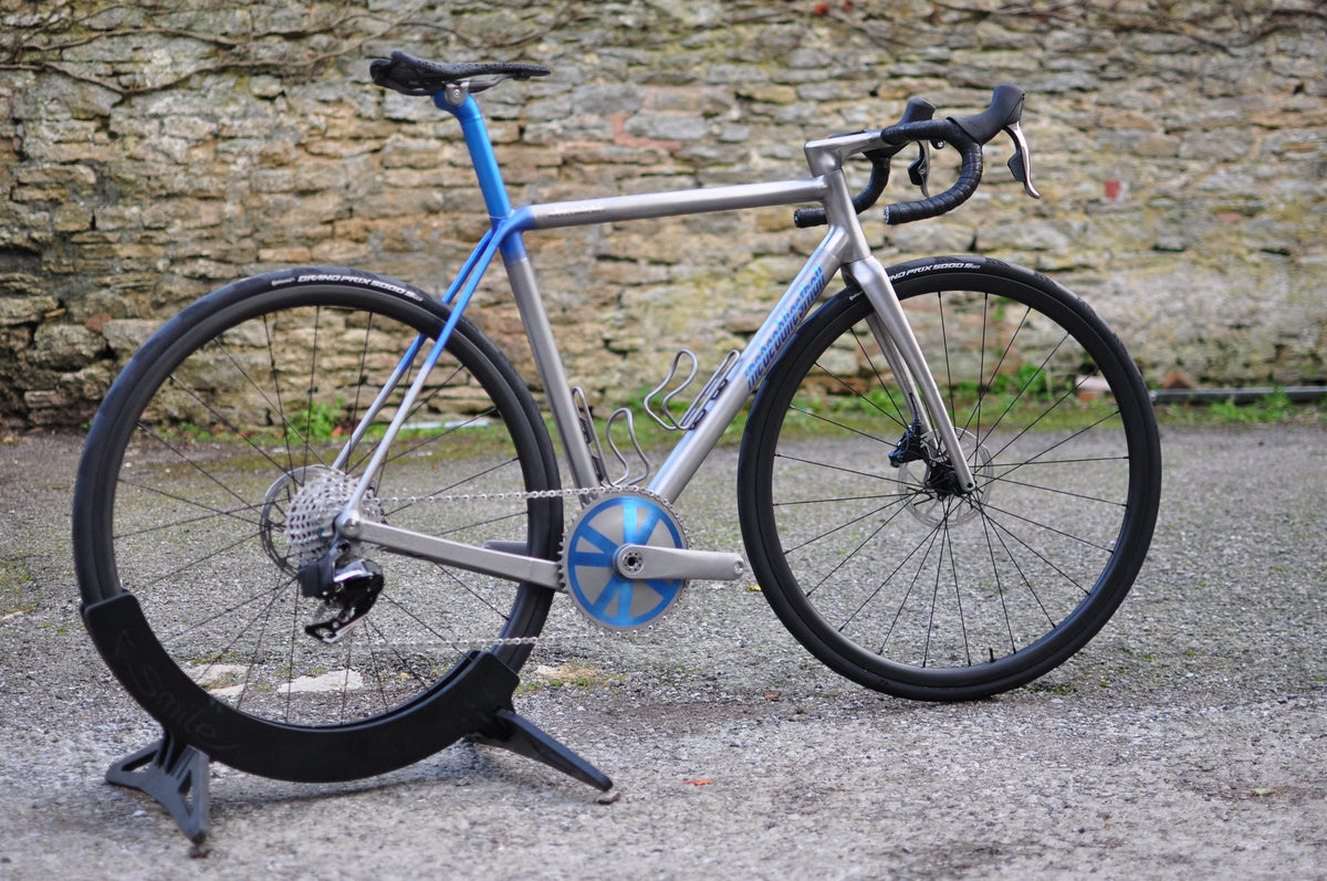 Titanium road bike with blue seat tube and stays, blue chainring cover and deep-section wheels by a stone wall