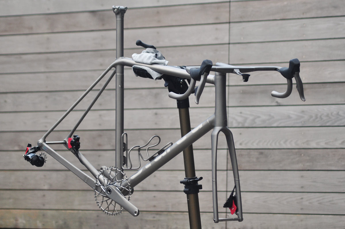 Bare titanium frame and fork part-built with handlebar and chainset on a workstand against timber cladding