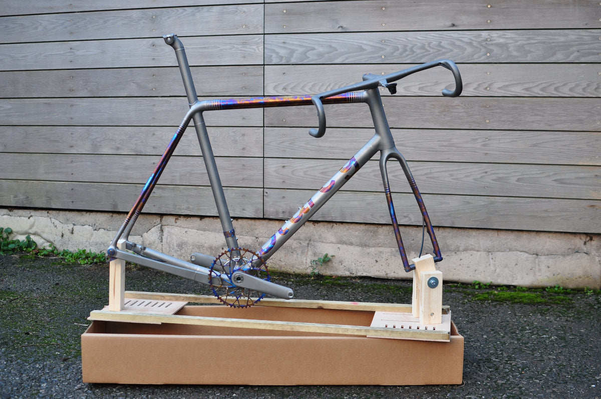 Titanium frame with rainbow anodised bands, part-built on a wooden packing jig in a cardboard box