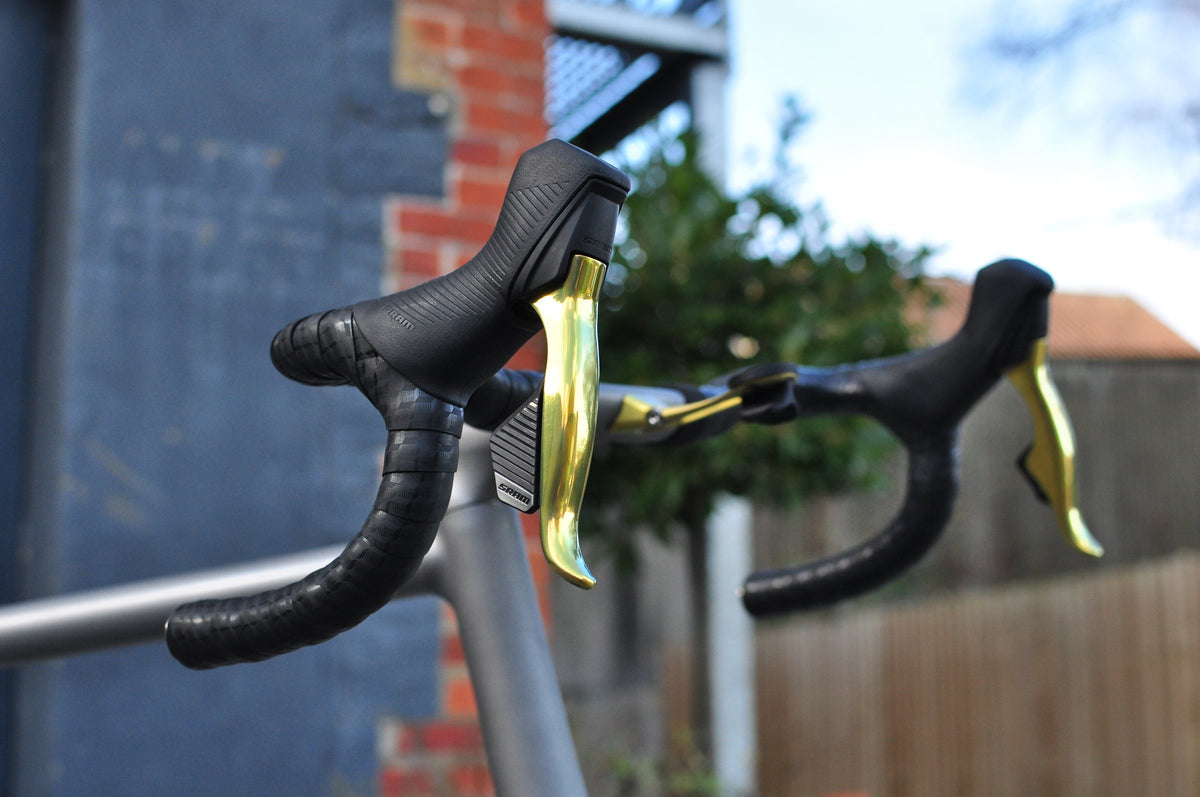 Drop handlebar with black hoods and gold brake levers on a titanium frame, brick wall behind