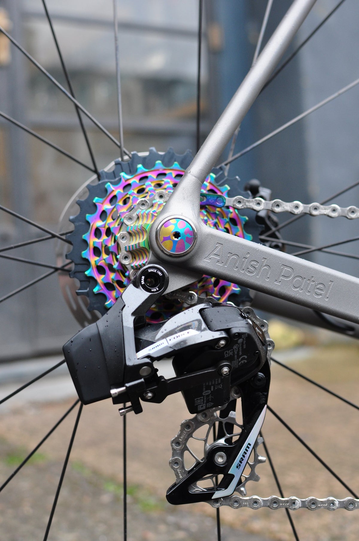 Titanium chainstay with engraved name, rainbow cassette and anodised axle bolt beside a rear mech
