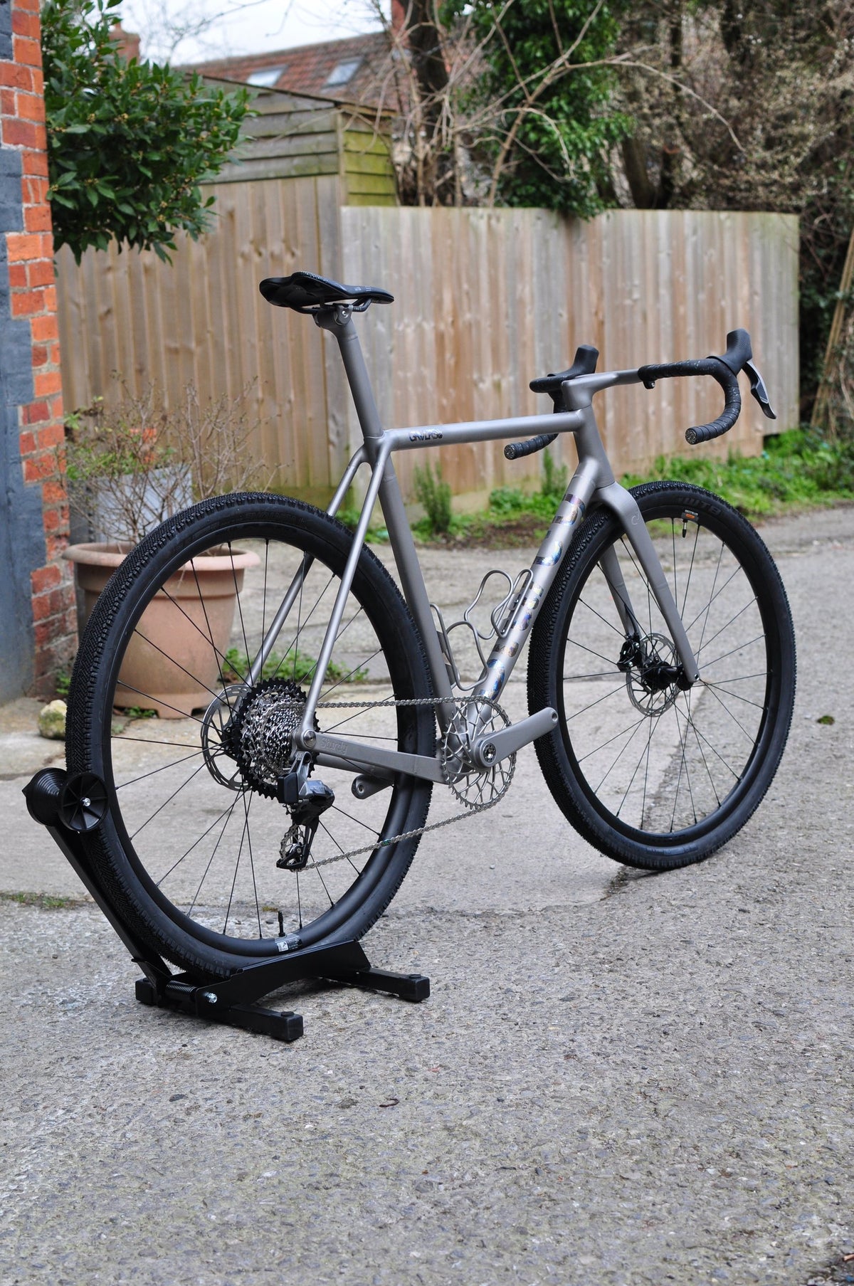 Titanium gravel bike with anodised down tube graphics and knobbly tyres on a stand by a garden fence