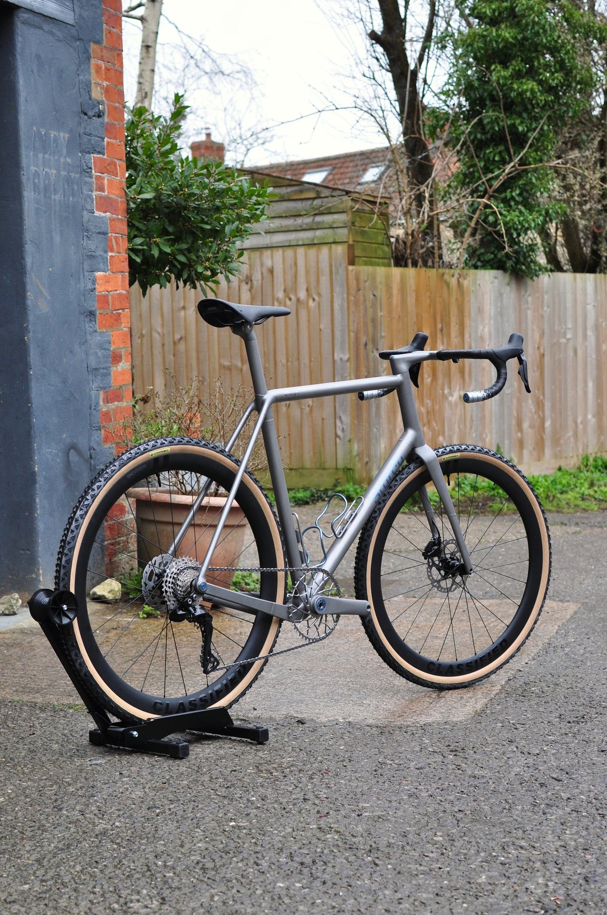 Titanium gravel bike with tan-wall tyres and blue anodised details on a stand by a garden fence