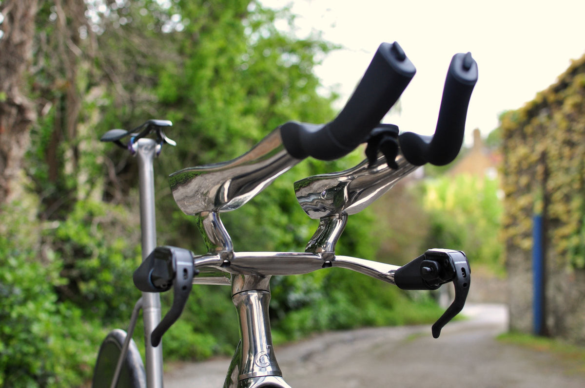 Polished titanium time trial base bar and extensions with black armrests in a leafy lane