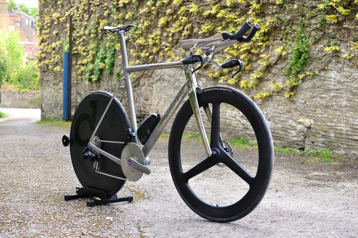 Titanium time trial bike with disc rear wheel, three-spoke front wheel and polished aero bars by an ivy wall