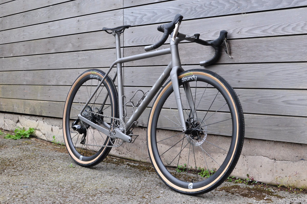 Titanium gravel bike with Sturdy wheels and tan-wall tyres leaning against grey timber cladding