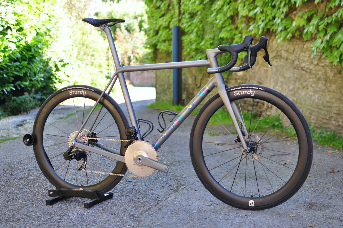 Titanium road bike with oil-slick anodised down tube logo and deep-section Sturdy wheels on a stand in a leafy lane