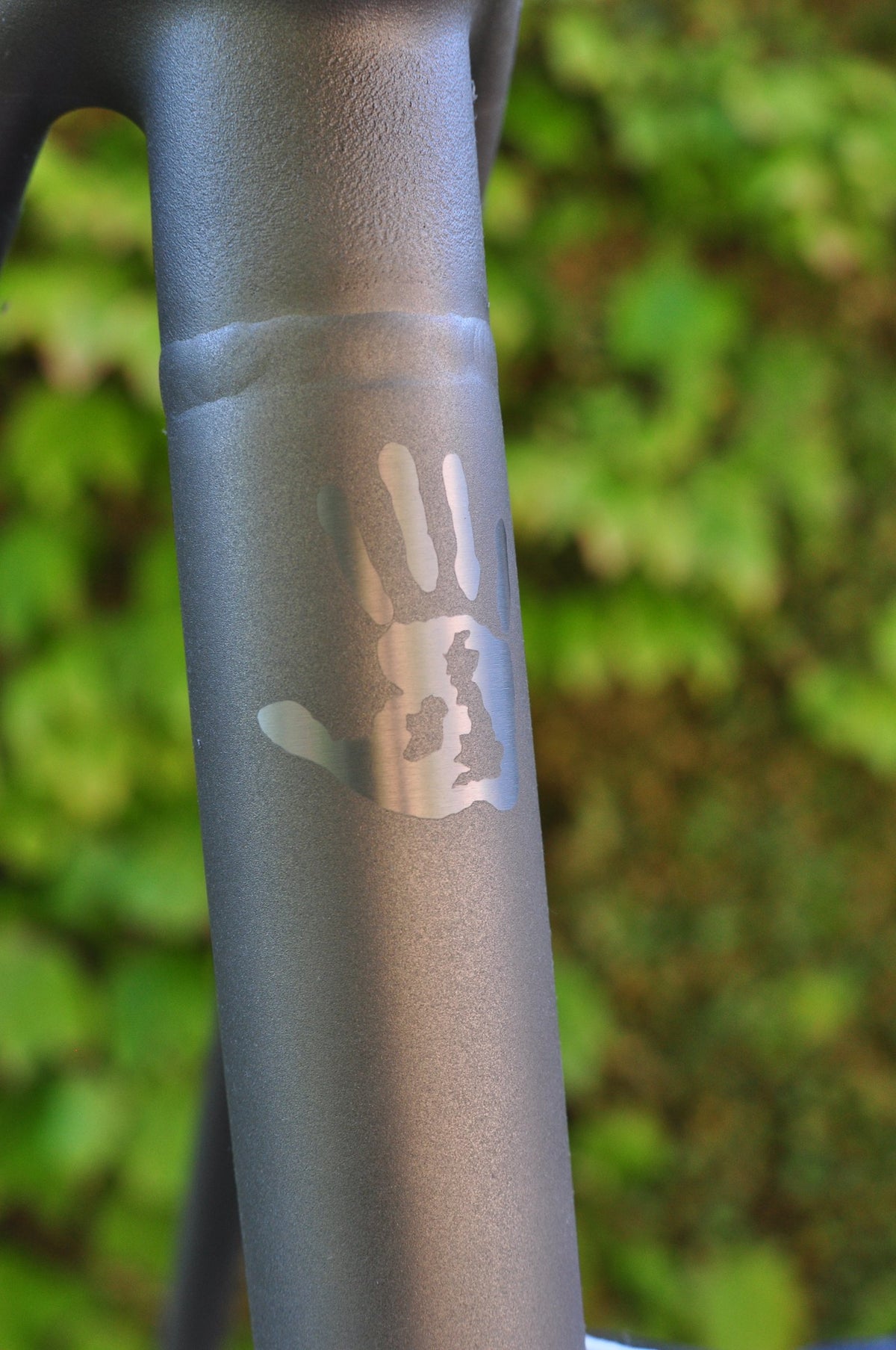 Polished handprint containing a map of Britain on a shot-peened titanium seat tube, greenery behind