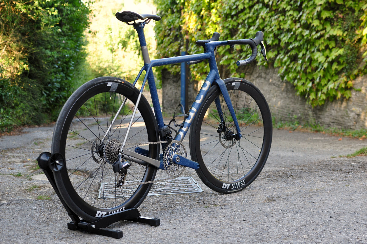 Road bike in navy speckled paint with bare titanium rear stays on a stand in a leafy lane