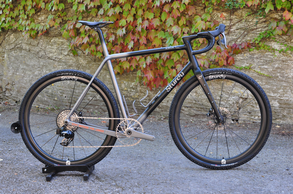 Titanium gravel bike with black fade and Sturdy logo, knobbly tyres, in a stand by an ivy-covered wall