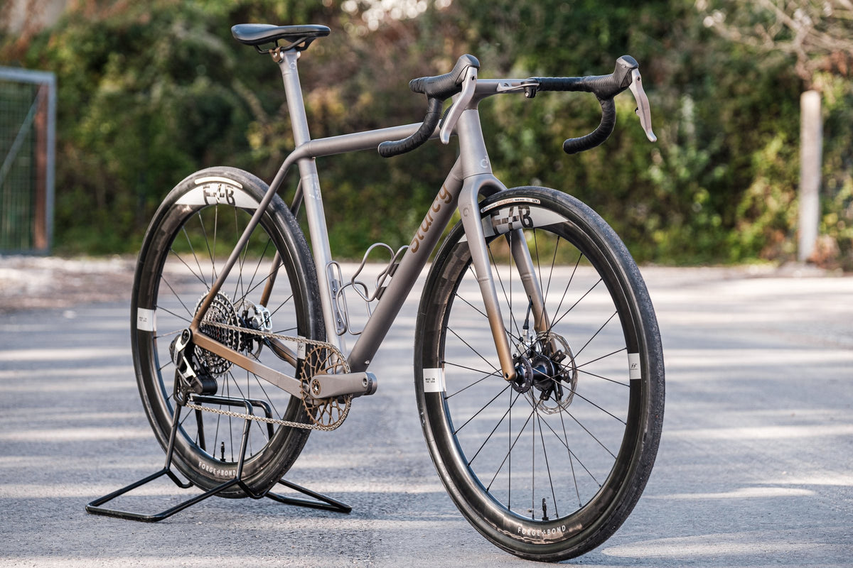 Brushed titanium gravel bike with bronze Sturdy lettering and deep carbon wheels on a sunlit road