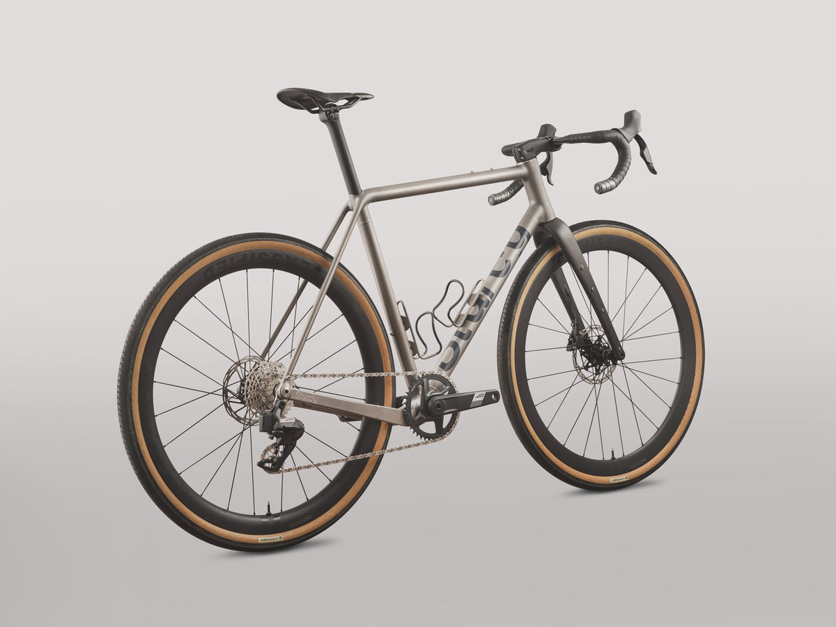 Titanium gravel bike with large grey down tube logo and tan-wall tyres against a plain grey backdrop