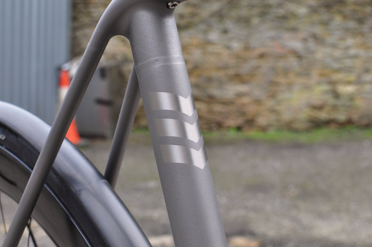 Shot-peened titanium seat tube with three polished chevron graphics, stone wall behind