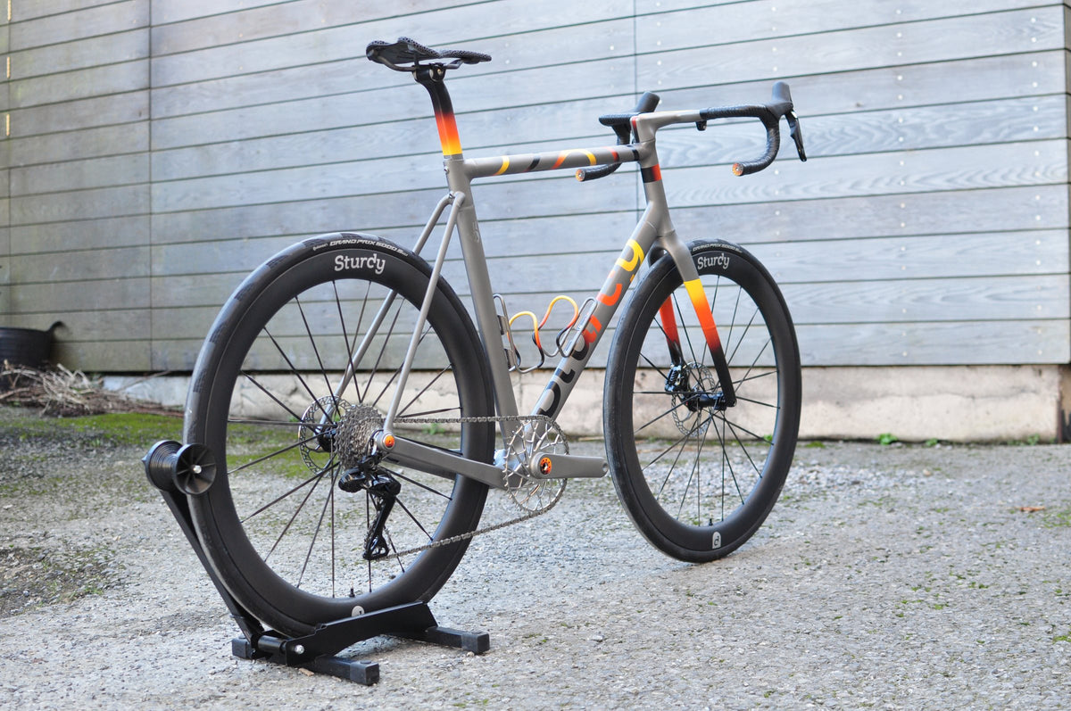 Titanium road bike with yellow, orange and red fade paint and Sturdy deep-section wheels on a stand by a timber wall