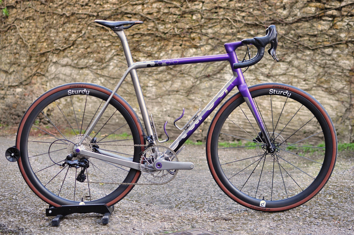 Titanium road bike with purple front end, hexagon fade on the top tube and tan-wall tyres by a stone wall