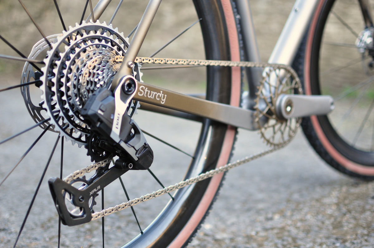 Titanium gravel bike drivetrain with wide-range cassette and Sturdy logo on the chainstay, close-up