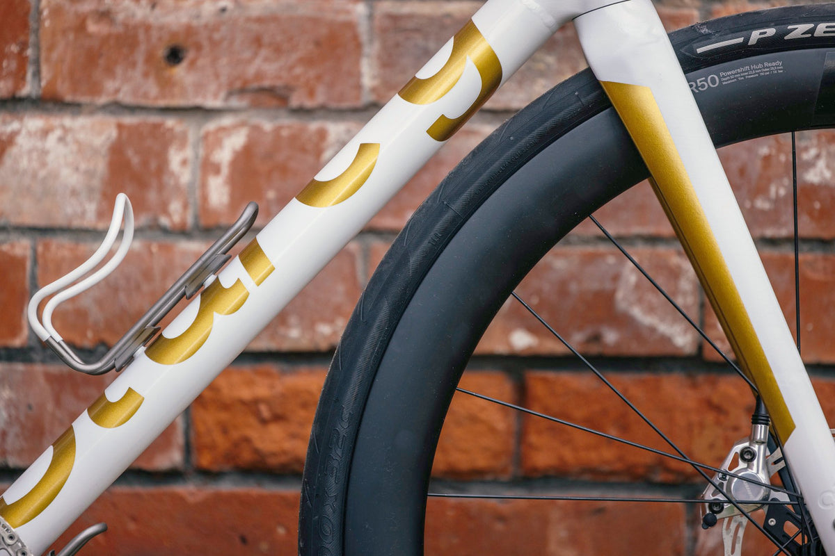 White down tube with gold lettering, white bottle cage and gold-striped fork against a red brick wall