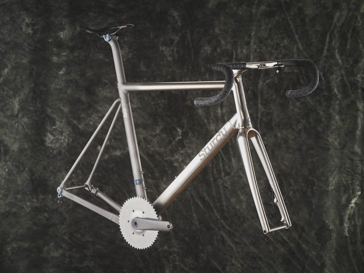 Bare titanium road frameset with Sturdy logo, black bar tape and single chainring against a dark mottled backdrop