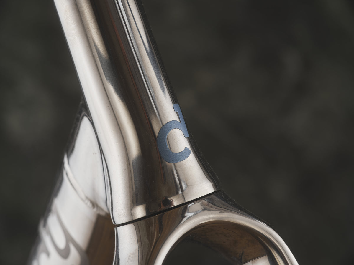 Polished titanium head tube junction with a small blue-grey logo, against a dark backdrop
