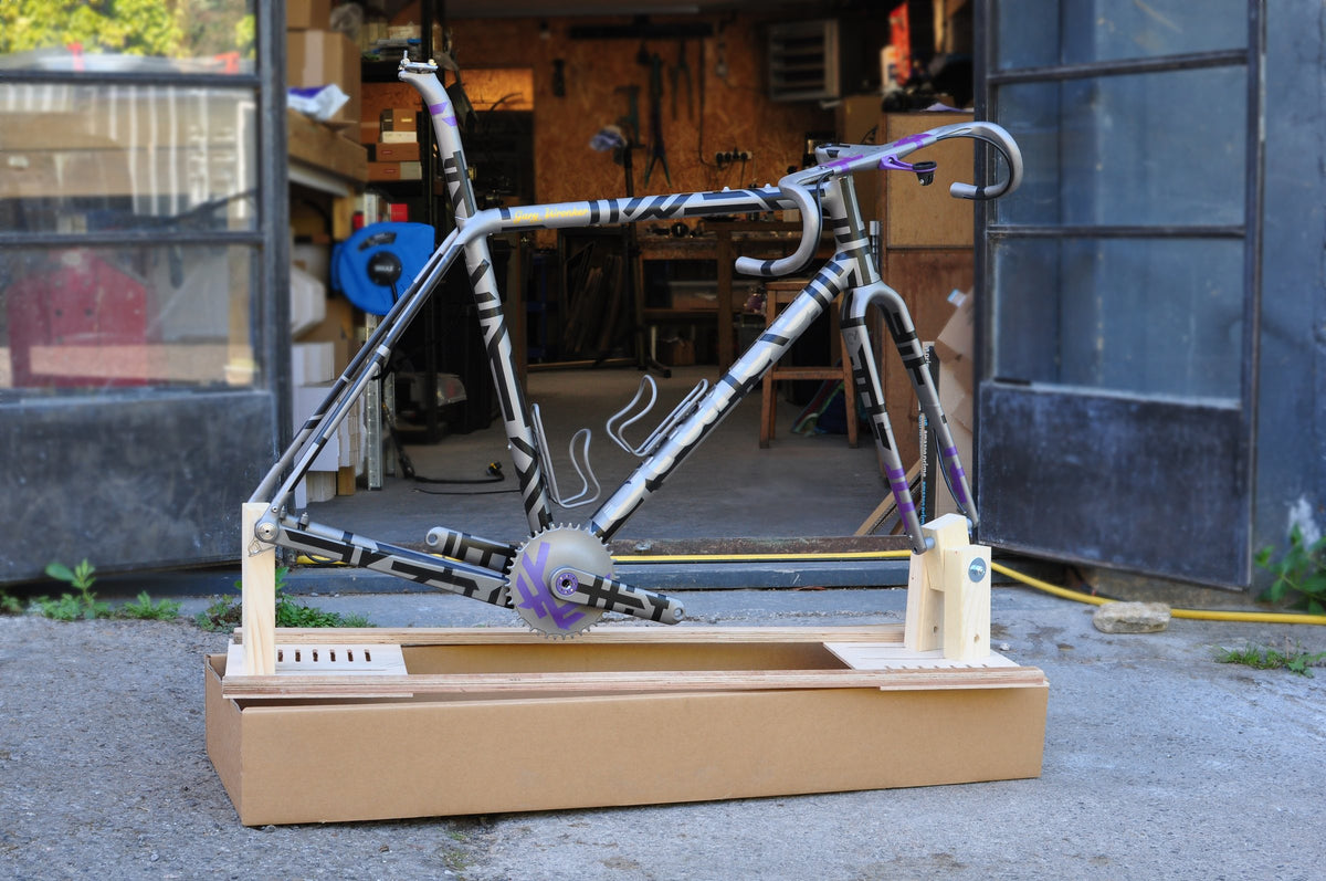 Titanium frameset with black and lilac geometric graphics on a wooden packing jig outside a workshop