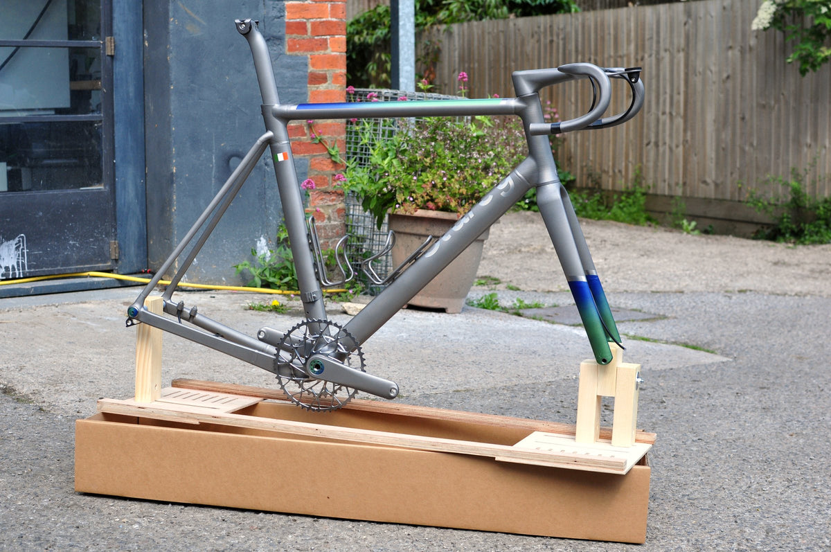 Titanium frameset with blue-to-green fade on the fork and top tube on a wooden packing jig in a yard