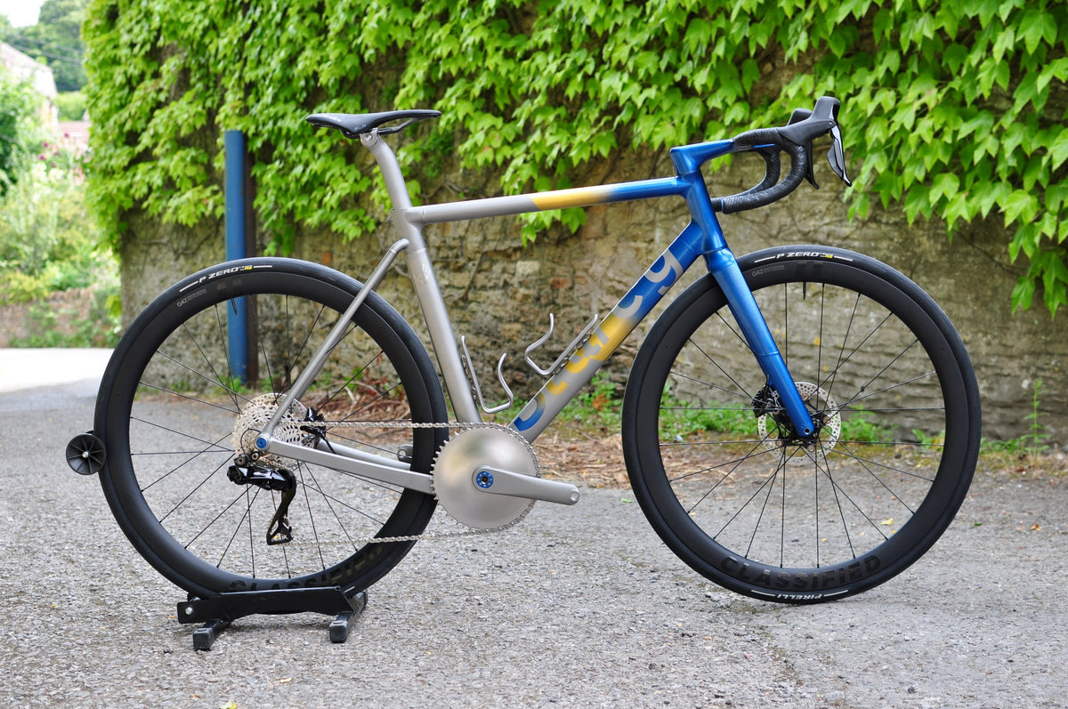 Titanium road bike with blue and gold fade front end and deep carbon wheels by an ivy-covered wall