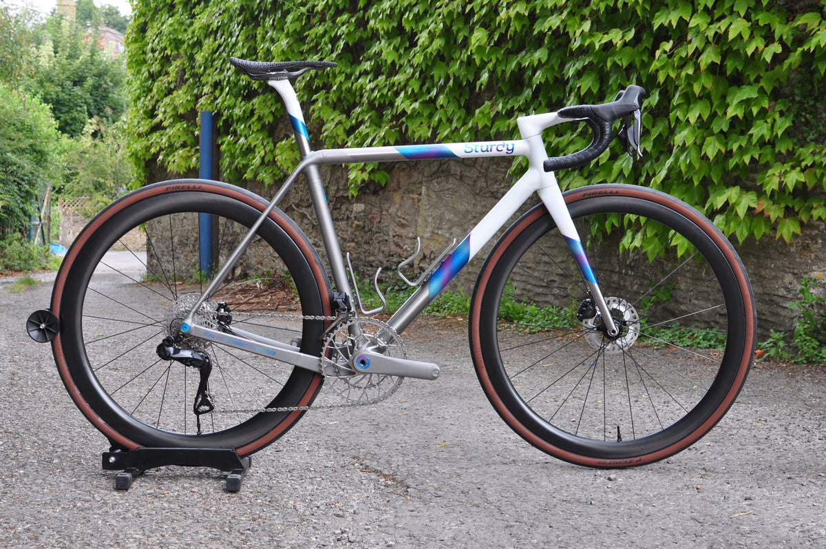 White and titanium road bike with blue-purple fade panels, Sturdy logo and tan-wall tyres on gravel