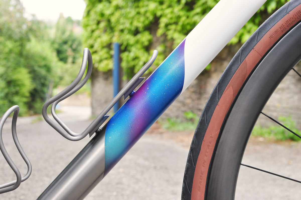 White and titanium down tube with sparkled blue-purple fade and titanium bottle cages, hedge behind