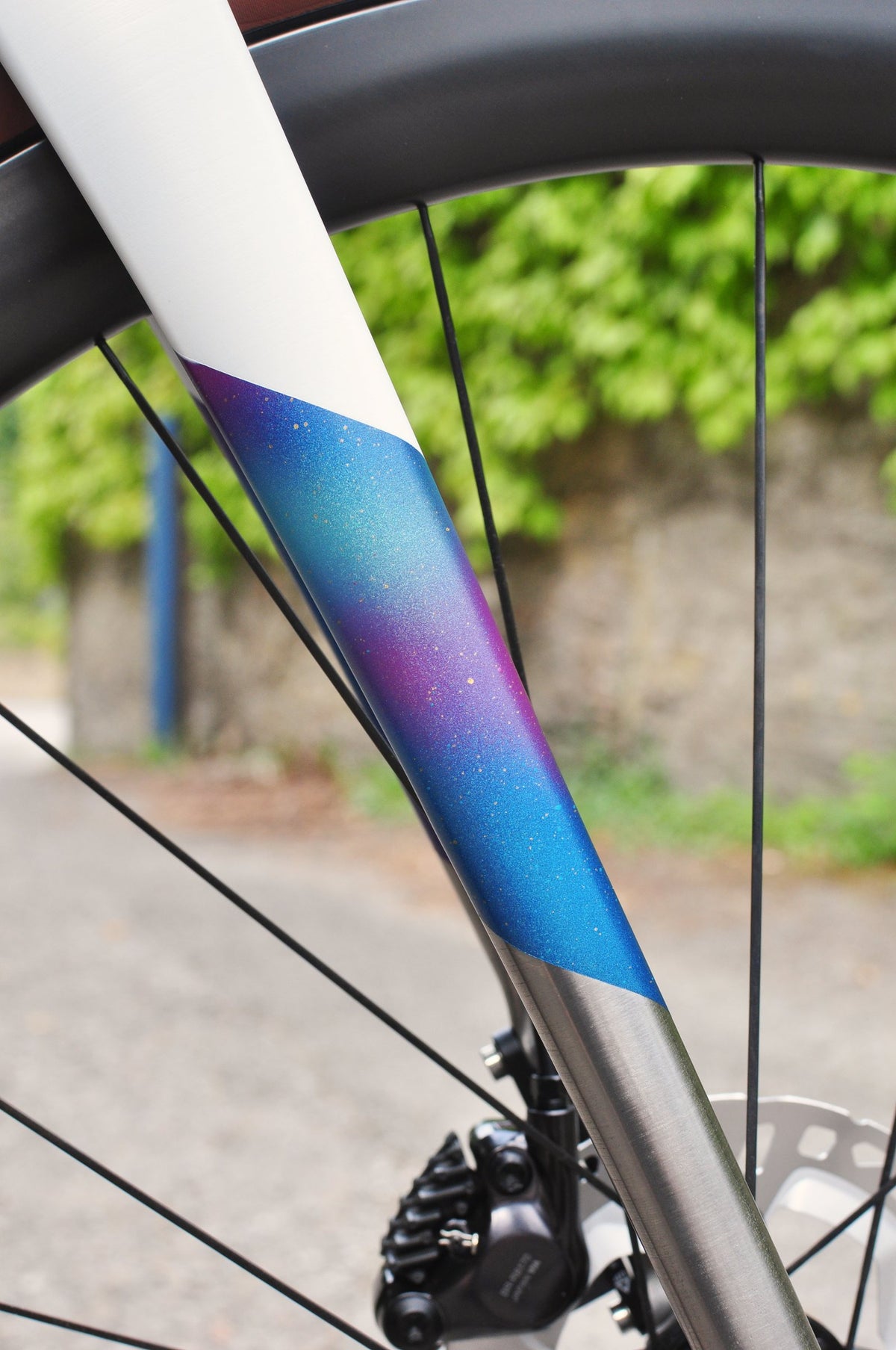 Fork blade in white with speckled blue-purple fade over brushed titanium, disc brake and spokes below